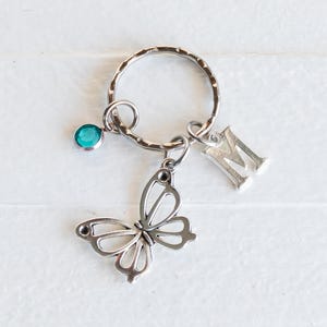 May include: Silver-toned keychain featuring a butterfly charm, a teal-colored gemstone, and an initial "M" pendant. The butterfly charm has an openwork design. The keychain is on a white surface.
