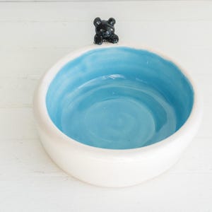 May include: A round ceramic bowl with a smooth white exterior and a bright sky blue interior. A small, sculpted black bear figure is attached to the top rim of the bowl, appearing to peer over the edge into the center of the basin.