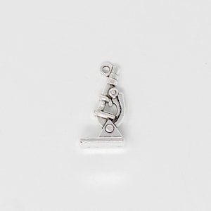 May include: A silver-colored microscope charm. The charm is a miniature replica of a microscope, featuring detailed components like the eyepiece, objective lenses, and base. It is a science-themed accessory.