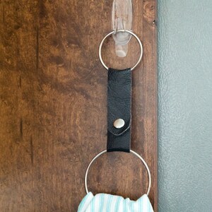 May include: A black leather towel ring with a silver button and a silver ring hanging from it. The ring is attached to a clear adhesive hook on a wooden surface.