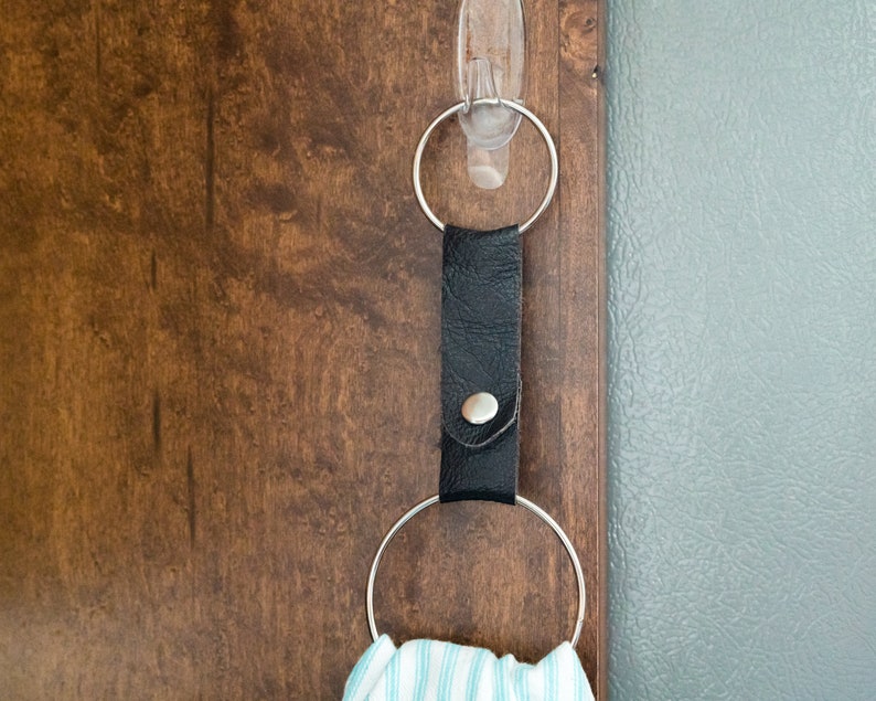 May include: A black leather towel ring with a silver button and a silver ring hanging from it. The ring is attached to a clear adhesive hook on a wooden surface.