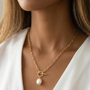 Puede incluir: Un collar de cadena dorada con un gran colgante de perla blanca. El collar presenta un cierre de palanca y un diseño sencillo y elegante. La cadena está hecha de eslabones rectangulares.