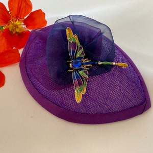 May include: A small purple hat with a blue mesh bow and a dragonfly brooch. The dragonfly is made of gold metal and has green and blue enamel accents.