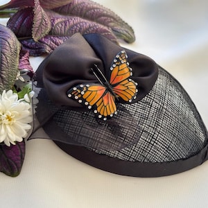May include: A black fascinator hat with a decorative orange and yellow butterfly. The butterfly has rhinestone accents. The hat has a black mesh base and a satin bow. The hat is next to purple leaves and a white flower.