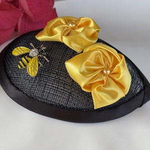May include: A black hat with a textured surface, adorned with two yellow satin bows and a bee-shaped brooch. The bee has yellow wings and a rhinestone body. The hat's brim is black.