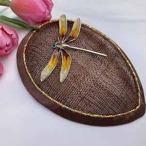 May include: A brown hat with a gold trim and a dragonfly brooch. The dragonfly is made of metal and has yellow, orange, and green accents.