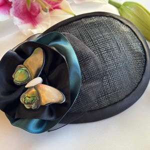 May include: A black fascinator with a textured, circular base and a decorative flower. The flower features black satin and teal ribbon, with iridescent abalone shell accents. A pink and white lily is in the background.