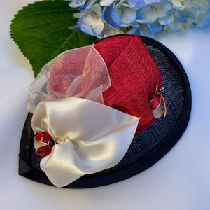 May include: A black, leaf-shaped fascinator with red and white fabric accents. The design includes a white satin bow and a red fabric section. Two ladybug brooches add a touch of whimsy. A sheer, light pink fabric is also included.