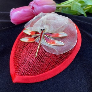 May include: A red and white fabric hat with a red dragonfly brooch. The hat is decorated with a white tulle bow.