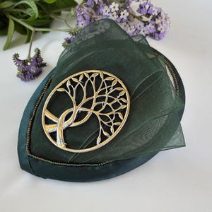 May include: A dark green fabric hat with a gold-coloured tree of life brooch. The hat has a leaf-like shape and is trimmed with a beaded edge. Purple flowers and green leaves are in the background.