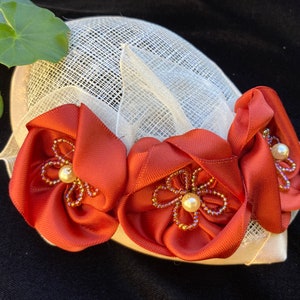 May include: A white mesh hat with three rust-colored fabric flowers adorned with pearl and gold beads.