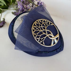 May include: A navy blue fascinator with a silver tree of life emblem. The hat features a sheer navy overlay and a beaded trim. The base is a mix of silver and navy fabric, creating a stylish accessory.