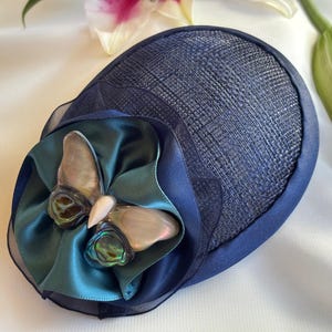 May include: A navy blue fascinator hat with a woven texture. The hat features a teal satin bow and a butterfly embellishment with iridescent wings. A white flower and green lily bud are in the background.
