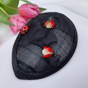 May include: A black fabric hat with three red ladybug embellishments. The hat is made of a textured fabric and has a black trim.