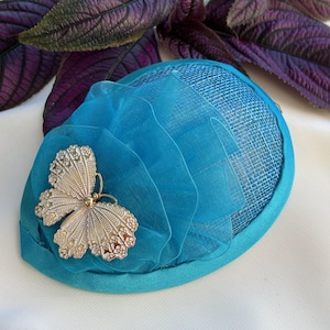 May include: A teal-colored fascinator with a silver butterfly embellishment. The headpiece features a mesh base, layered sheer fabric, and a satin trim. The butterfly has intricate detailing and a gold-colored center.