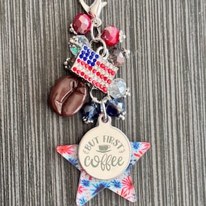 May include: A silver charm with a lobster clasp, featuring red and blue beads, a rhinestone American flag, and a coffee bean. A star-shaped charm with the text "But First Coffee" completes the design.