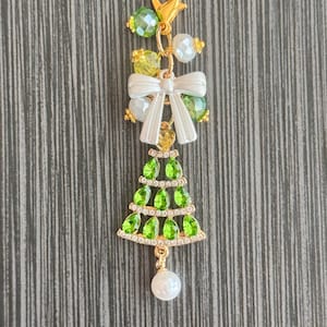 May include: A gold-colored charm with a Christmas tree design. The charm features green and clear rhinestones, a white bow, and green and white beads. A white pearl hangs from the bottom. The charm is attached to a gold-colored clasp.
