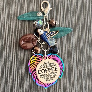 May include: A coffee-themed charm with a lobster clasp. It features a coffee bean, a blue bird, and a leaf-shaped charm with the word "COFFEE" and coffee-related terms. The charm has iridescent edges and is accented with beads.