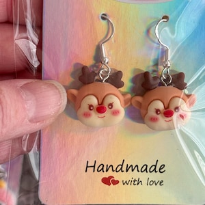 May include: A pair of reindeer earrings with silver hooks. The reindeer have brown antlers, tan faces, red noses, and pink cheeks. The earrings are displayed on a card that reads "Bowtique Charms" and "Handmade with love."