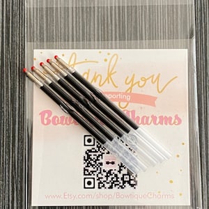 May include: A close-up of six black ink refills with red tips, packaged in a clear plastic bag. The bag has a white card with the text "Thank you for supporting Bowtique Charms" and a QR code.