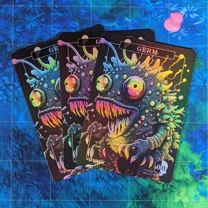 May include: Three holographic trading cards featuring a colorful, grotesque, cartoon monster with large eyes and sharp teeth. The text on the card reads "GERM".