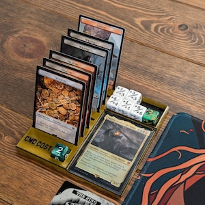 May include: A wooden game organizer with a yellow and brown color scheme. The organizer has a slot for cards and a compartment for dice. The organizer has the text "CMD COST" printed on it. There are several cards in the organizer, including a card with the text "The Treasure" and a card with the text "Sauro, Lord of the Rings". There are also several dice in the compartment, including a green die with a black "6" on it.