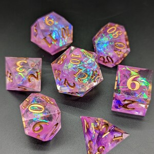 May include: Set of seven iridescent purple dice with gold numbers. The dice are made of clear resin with a swirling purple and iridescent effect. The numbers are in gold and are visible on each face of the dice.