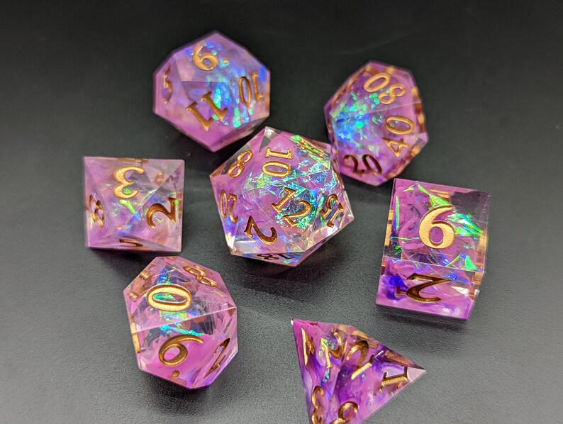 May include: Set of seven iridescent purple dice with gold numbers. The dice are made of clear resin with a swirling purple and iridescent effect. The numbers are in gold and are visible on each face of the dice.