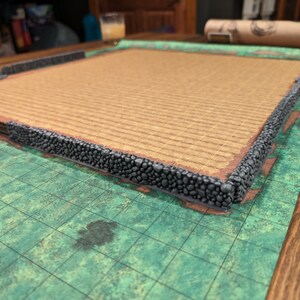 Puede incluir: Una pieza de terreno para juegos de mesa con un borde de muro de piedra. El interior tiene una superficie texturizada de color marr&oacute;n claro. La pieza se encuentra sobre un mapa de cuadr&iacute;cula verde y marr&oacute;n, con una mesa de madera en el fondo.