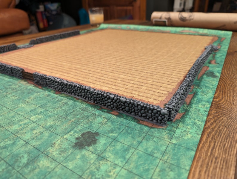 Puede incluir: Una pieza de terreno para juegos de mesa con un borde de muro de piedra. El interior tiene una superficie texturizada de color marr&oacute;n claro. La pieza se encuentra sobre un mapa de cuadr&iacute;cula verde y marr&oacute;n, con una mesa de madera en el fondo.