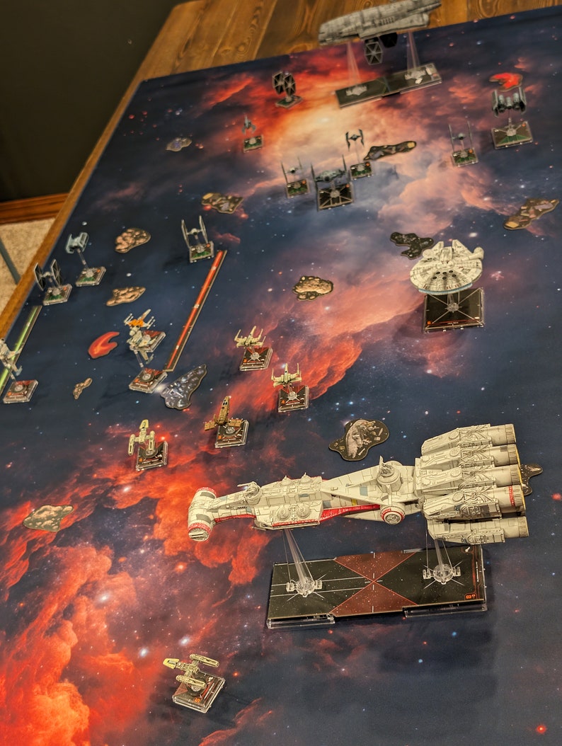 May include: A close-up view of a Star Wars X-Wing miniature spaceship game set up on a game board with a nebula background. The game pieces are arranged in a strategic formation, with a large spaceship in the foreground.