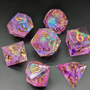May include: Set of seven iridescent purple dice with gold numbers. The dice are made of clear resin with a glittery finish. The dice are in various shapes, including a d4, d6, d8, d10, d%, d12, and d20.