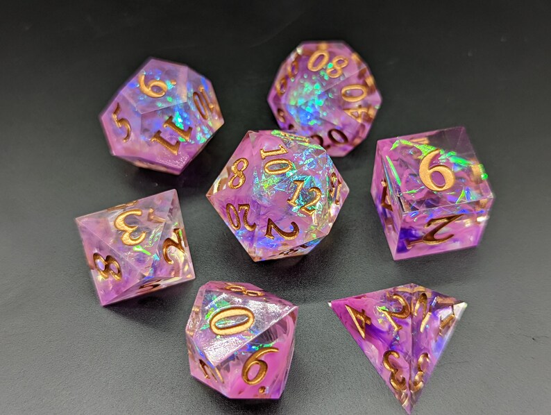 May include: Set of seven iridescent purple dice with gold numbers. The dice are made of clear resin with a glittery finish. The dice are in various shapes, including a d4, d6, d8, d10, d%, d12, and d20.