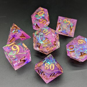 May include: Set of seven iridescent pink and purple dice with gold numbers. The dice are made of a clear resin with a swirling pattern of pink and purple.