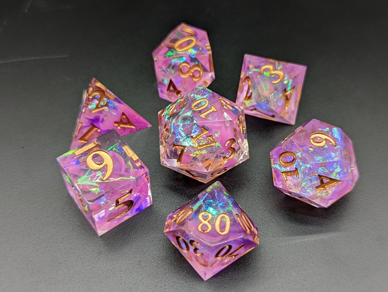 May include: Set of seven iridescent pink and purple dice with gold numbers. The dice are made of a clear resin with a swirling pattern of pink and purple.