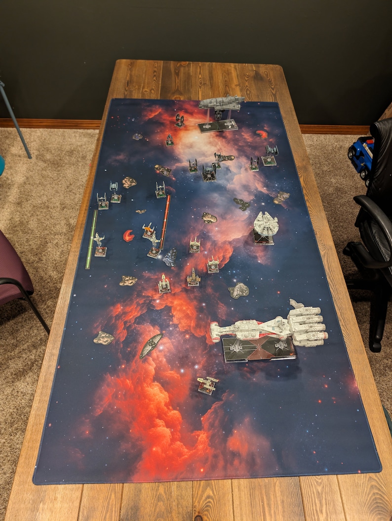 May include: A close-up view of a game board with a space-themed design. The board features a dark blue background with red and orange nebulae. There are several miniature spaceships and other game pieces scattered across the board.