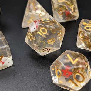 May include: Set of clear dice with gold numbers and a white fish design inside. The dice are on a black surface.