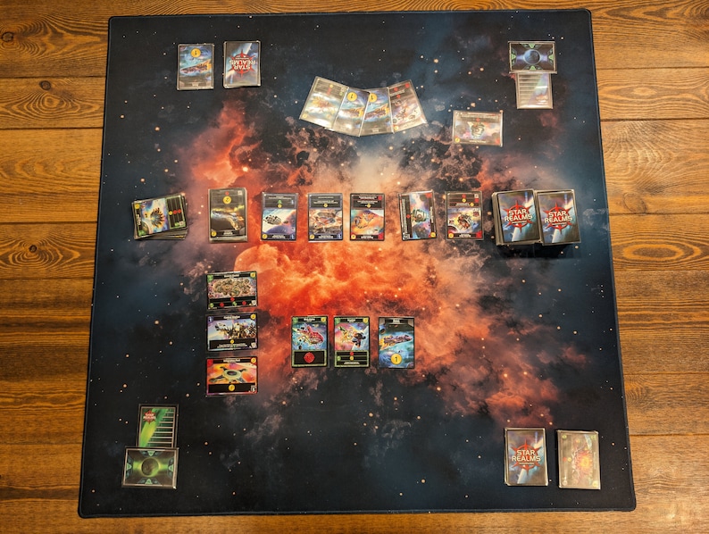 May include: A game board with a nebula background and cards laid out for a game of Star Realms. The cards are blue, red, and black with various images and text. There are two decks of cards labeled "Star Realms" on the table.