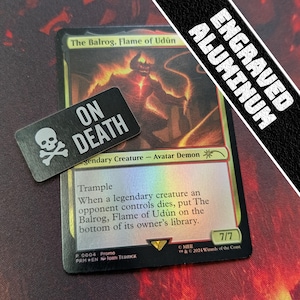 Puede incluir: Una carta de Magic: The Gathering con "The Balrog, Flame of Udûn" y un marcador negro "On Death". La carta tiene un acabado holográfico y una fuerza/resistencia de 7/7. El marcador tiene una calavera y huesos cruzados.
