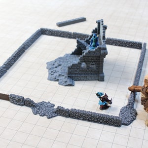 Puede incluir: Una escena de juego de mesa de fantas&iacute;a en miniatura con un recinto de muro de piedra, una estructura de castillo en ruinas y varias figuras. La escena incluye una figura grande y musculosa y una figura m&aacute;s peque&ntilde;a con detalles azules. Las paredes son grises y las figuras est&aacute;n pintadas.