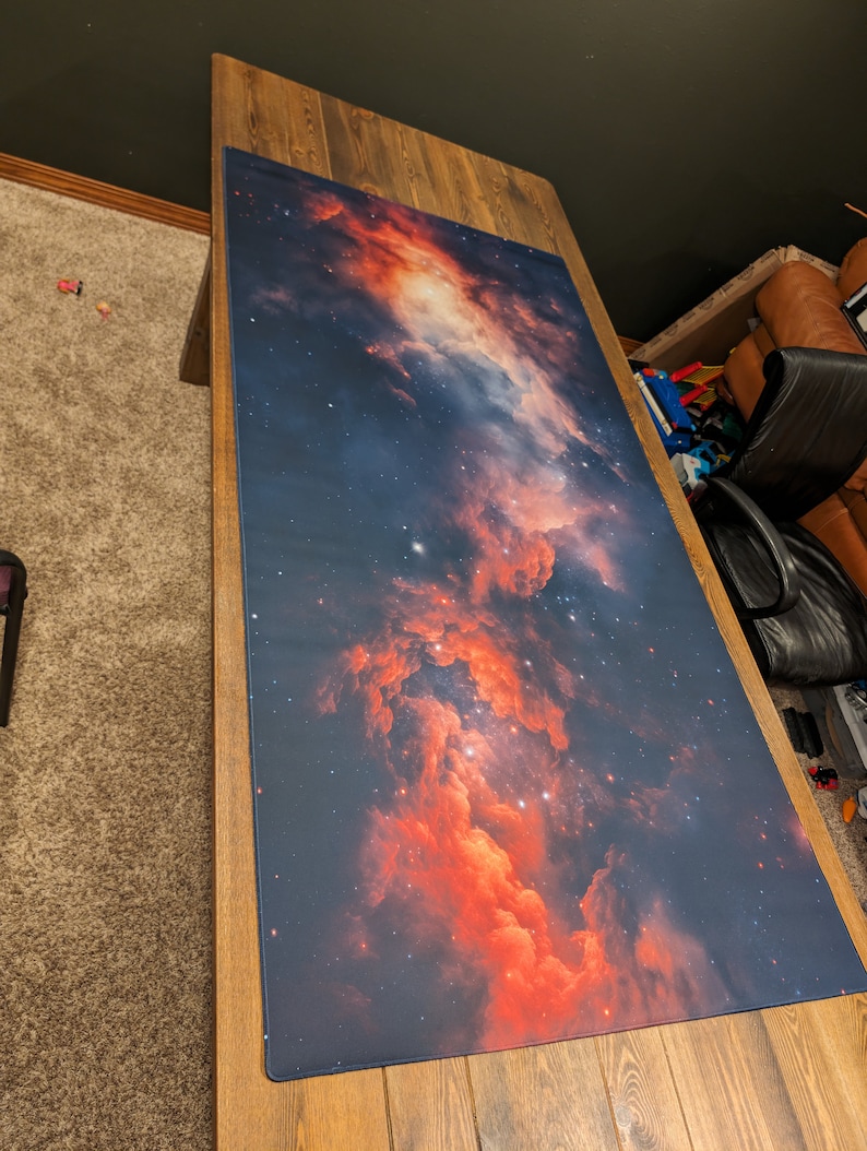 May include: A large, rectangular, dark blue gaming mat with a red and orange nebula design. The mat is laid out on a wooden table.