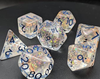 Op de afbeelding: Set van zeven iriserende dobbelstenen met blauwe nummers. De dobbelstenen zijn helder en hebben een glinsterende afwerking. De nummers op de dobbelstenen zijn 1, 2, 3, 4, 5, 6, 7, 8, 9, 10, 11 en 12.