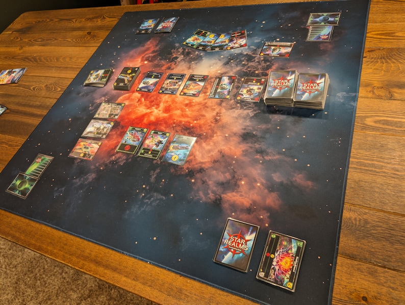 May include: A game board with a nebula background and several cards laid out. The cards are from the Star Realms game. There are two stacks of cards labeled "Star Realms" on the board.