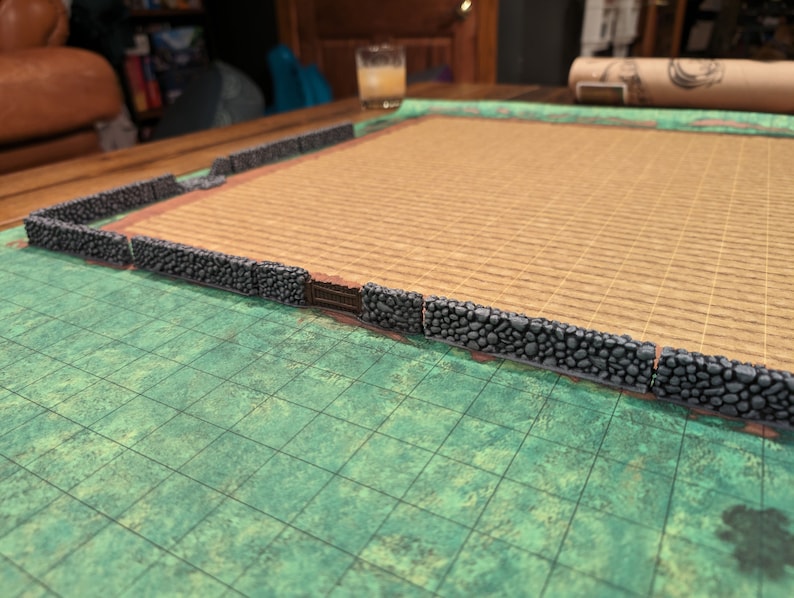Puede incluir: Un muro de piedra en miniatura con una puerta rodea una esterilla beige, colocada sobre una superficie de rejilla verde. El muro est&aacute; hecho de piedras grises texturizadas. La esterilla tiene un aspecto tejido, y la rejilla es de un verde m&aacute;s oscuro.