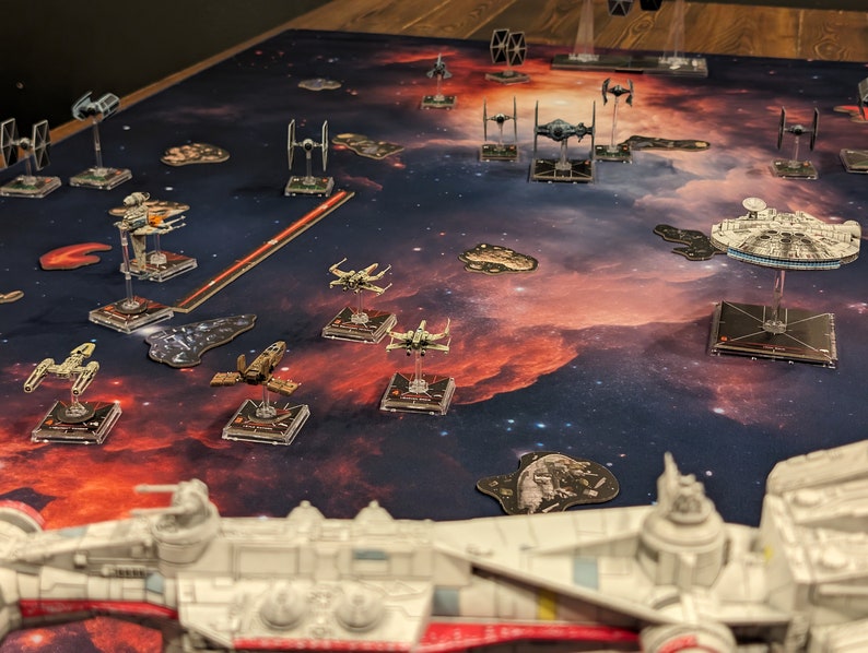 May include: A close-up of a Star Wars X-Wing miniature spaceship game set up on a game board with a nebula background. The game board features a variety of spaceships, including X-Wings, TIE fighters, and a Millennium Falcon.