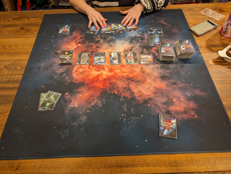 May include: A game board with a nebula design, featuring a deck of cards with the text "Star Realms" on the top card. The board is laid out on a wooden table with a stack of cards and a cell phone.