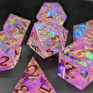 May include: Set of seven iridescent pink dice with gold numbers. The dice are made of a clear material with a pink and iridescent finish. The numbers are in gold and are easy to read.