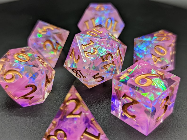 May include: Set of seven iridescent pink dice with gold numbers. The dice are made of a clear material with a pink and iridescent finish. The numbers are in gold and are easy to read.