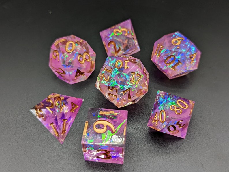 May include: Set of seven iridescent purple and gold dice with a variety of shapes and numbers. The dice have a glittery, holographic finish.