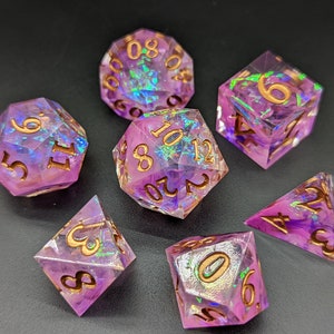 May include: Set of seven iridescent purple dice with gold numbers. The dice are made of clear resin with a purple tint and have a glittery finish. The numbers are printed in gold.